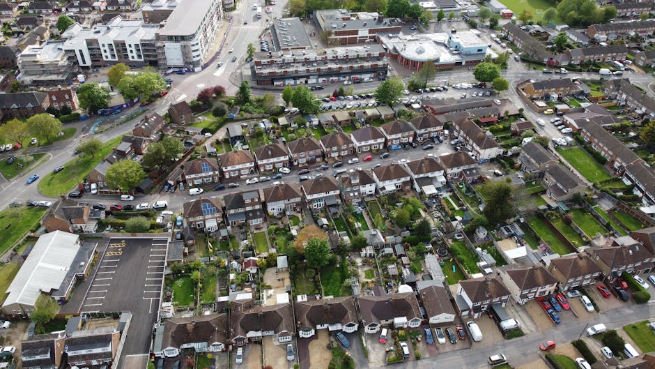 An aerial view of a residential neighborhood featuring a series of terraced houses with small, well-maintained gardens. The gardens display a variety of textures including neatly trimmed grass lawns,